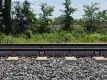 Railway tracks with L.B. Foster branded strain gauge sensors mounted on the rails between concrete sleepers, surrounded by ballast stones, with lush green vegetation in the background.